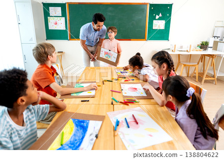 The teacher stand beside and try to make a little boy with blond hair and wear glasses present his drawing or painting in front of the classroom and also ask the idea or the meaning of the picture. 138804622