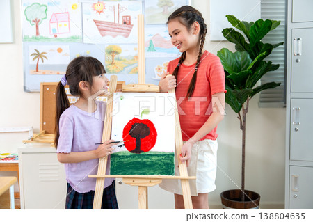 Portrait of mix-raced two girls stand beside of their painting that they create together in the classroom and they look each other with smiling. Portrait of mix-raced two girls stand beside of their painting that they create together in the classroom and they look each other with smiling. 138804635