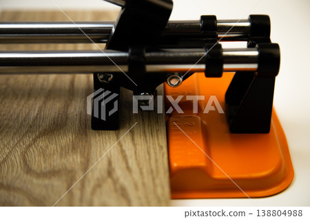 Close-up side view of an orange mechanical tile cutter, which fixed ceramic tiles of wooden colors, above it is a cutting disc on a white background. The concept of renovation, tiling, construction 138804988