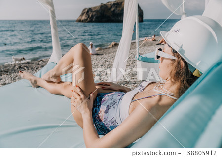 Beach woman hardhat relaxes on a sunbed wearing a floral swimsuit and safety helmet on a beautiful pebble shore 138805912