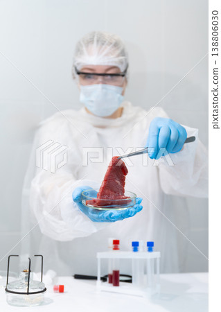 Woman scientist in protective gear handling lab-grown meat sample with tongs in petri dish. Artificial meat cultivation in biotechnology laboratory. Sustainable protein production and innovative food 138806030