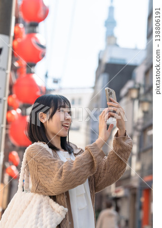 一位微笑的女子一邊用智慧型手機拍照，一邊漫步在市中心，遠處可以看到東京晴空塔。 138806191