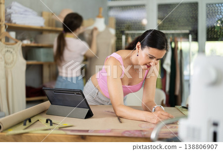 Woman tailor draws dress pattern of product on paper, tablet on table. Companion in background 138806907
