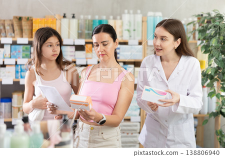 Attentive young female pharmacist advising family of mother and daughter choosing box with pills in pharmacy store 138806940