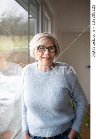 Smiling elderly woman looking through window at home. Smiling elderly woman looking through window at home. 138809122