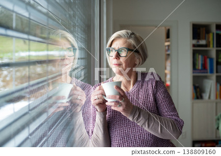 Elderly woman having cup of tea and looking through window at home. Elderly woman having cup of tea and looking through window at home. 138809160