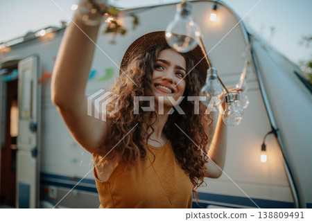 Young beautiful woman is on a camping trip in nature, putting lights on tree, preparing for evening. 138809491