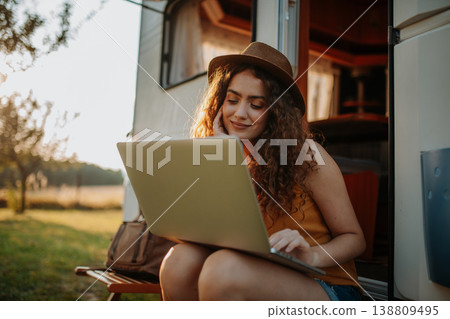 Young beautiful woman is on a camping trip in nature, sitting in front of caravan with notebook on knees. Caravan as homeoffice, working remotely. 138809495