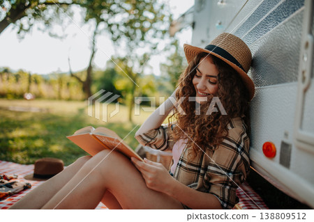 Young beautiful woman is on a camping trip in nature, sitting in front caravan and reading book. Young beautiful woman is on a camping trip in nature, sitting in front caravan and reading book. 138809512