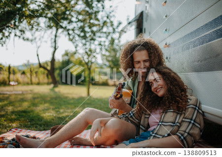 Couple sitting by caravan, enjoying peaceful moment. Man playing guitar and singing song to his girlfriend. Couple sitting by caravan, enjoying peaceful moment. Man playing guitar and singing song to his girlfriend. 138809513