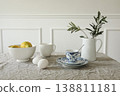 Minimalist breakfast still life. Lemons, eggs, coffee cup olive branch in white jug on linen table. Scandinavian kitchen scene. Natural light, neutral tones. Modern Mediterranean rustic home styling. 138811181