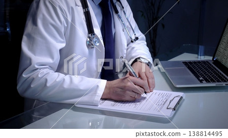 Physician writing patient medical history notes on a clipboard while sitting at a desk with a laptop, managing documents and providing healthcare services in a clinic or office Physician writing patient medical history notes on a clipboard while sitting at a desk with a laptop, managing documents and providing healthcare services in a clinic or office 138814495