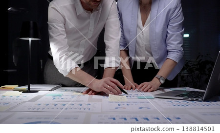 Business colleagues collaborating late at night, hands pointing at financial documents with graphs and charts, developing strategy in a dimly lit office Business colleagues collaborating late at night, hands pointing at financial documents with graphs and charts, developing strategy in a dimly lit office 138814501