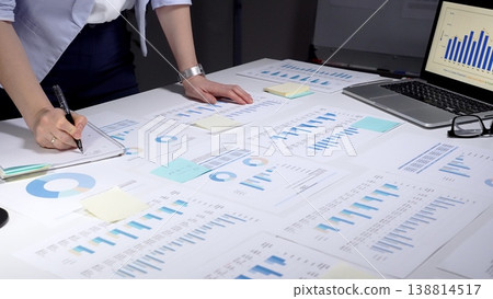 Business professional analyzing financial data with charts and graphs, working late in an office preparing reports and organizing information with sticky notes and a pen Business professional analyzing financial data with charts and graphs, working late in an office preparing reports and organizing information with sticky notes and a pen 138814517
