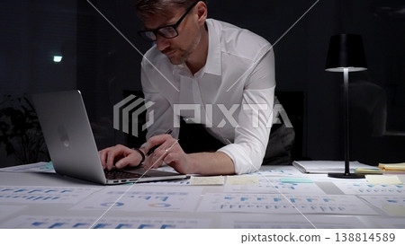 Businessman working late, leaning over a desk of financial reports and charts, typing on a laptop under a desk lamp, focused on analysis and planning into the night Businessman working late, leaning over a desk of financial reports and charts, typing on a laptop under a desk lamp, focused on analysis and planning into the night 138814589