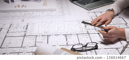 Architect carefully drawing detailed blueprints for a new construction project at a cluttered desk with laptop, scale model, ruler and pens, showing precision and professional focus, close up 138814667
