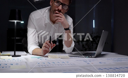 Dedicated businessman intensely analyzing financial documents and charts spread across his desk under a table lamp, concentrating on complex data in a dark office while working late evenings Dedicated businessman intensely analyzing financial documents and charts spread across his desk under a table lamp, concentrating on complex data in a dark office while working late evenings 138814720