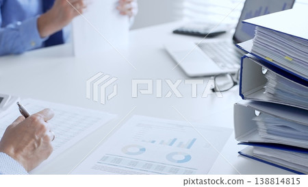 Corporate professionals reviewing financial documents, working collaboratively at white desk with blue folder stack, highlighting teamwork and business productivity. Audit and taxes in business 138814815