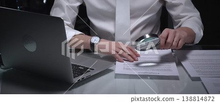 Businessman examining a document using a magnifying glass, focusing on fine print and terms in a legal agreement, working late. Audit and overworking concepts 138814872