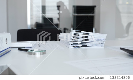 Office desk with stack of paper and magnifying glass, symbolizing heavy bureaucracy and business workload. Audit and taxes in business Office desk with stack of paper and magnifying glass, symbolizing heavy bureaucracy and business workload. Audit and taxes in business 138814896