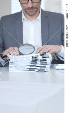 A businessman at his office desk, handling financial calculations, managing accounts, and reviewing documents, holding a magnifying glass. Audit and taxes in business 138814923