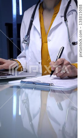 Doctor writing patient medical records, managing data, and documenting information on a clipboard in a clinic setting, utilizing modern technology with a laptop Doctor writing patient medical records, managing data, and documenting information on a clipboard in a clinic setting, utilizing modern technology with a laptop 138815088
