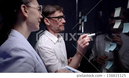Business professionals collaborating on a strategic plan. Analyzing data on a digital tablet in a dark modern office with sticky notes on a glass board Business professionals collaborating on a strategic plan. Analyzing data on a digital tablet in a dark modern office with sticky notes on a glass board 138815125