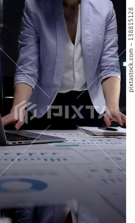 Businesswoman analyzing corporate financial data and sales growth charts on documents spread across an office desk, working late night in a professional setting 138815128