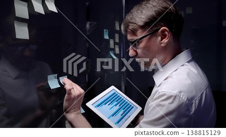 Business professional analyzing data on a tablet and planning strategy with adhesive notes on a glass board, brainstorming and organizing ideas for project development in a modern office 138815129