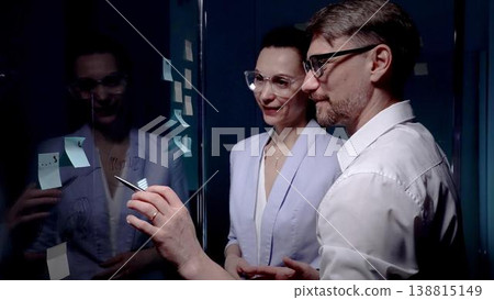 Business professionals collaborating around a glass board with sticky notes, brainstorming strategies and plans, writing ideas and analyzing data in a modern office team meeting Business professionals collaborating around a glass board with sticky notes, brainstorming strategies and plans, writing ideas and analyzing data in a modern office team meeting 138815149