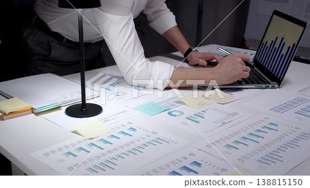 Professional businessman analyzing financial data and charts on laptop and paper documents, writing notes late at night, working overtime in modern office researching company growth and strategy Professional businessman analyzing financial data and charts on laptop and paper documents, writing notes late at night, working overtime in modern office researching company growth and strategy 138815150
