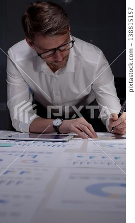 Businessman leans over a desk strewn with charts and reports, working on a tablet in a dim office late at night, reflecting overtime, focus, and relentless dedication Businessman leans over a desk strewn with charts and reports, working on a tablet in a dim office late at night, reflecting overtime, focus, and relentless dedication 138815157