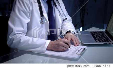 Physician writing patient medical history notes on a clipboard while sitting at a desk with a laptop, managing documents and providing healthcare services in a clinic or office Physician writing patient medical history notes on a clipboard while sitting at a desk with a laptop, managing documents and providing healthcare services in a clinic or office 138815164