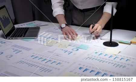 Business professional studies financial reports and market data at a cluttered desk, surrounded by charts, graphs and a laptop, focused on planning, strategy and results Business professional studies financial reports and market data at a cluttered desk, surrounded by charts, graphs and a laptop, focused on planning, strategy and results 138815167
