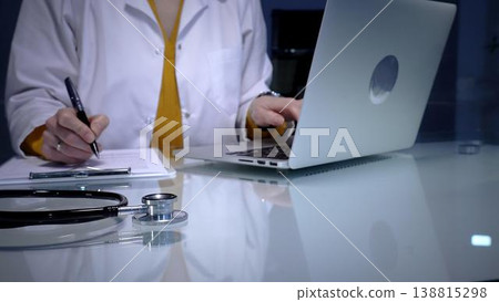 Stethoscope resting on a white reflective desk while medical professional working in clinic office. Wearing lab coat. Typing on laptop and writing notes. Stethoscope resting on a white reflective desk while medical professional working in clinic office. Wearing lab coat. Typing on laptop and writing notes. 138815298