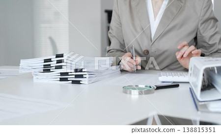 Businesswoman reviewing financial reports, calculating taxes, and managing audit paperwork in a busy office setting. Audit and taxes concept Businesswoman reviewing financial reports, calculating taxes, and managing audit paperwork in a busy office setting. Audit and taxes concept 138815351