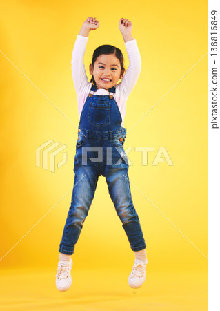 Portrait, kids and jump with a winner girl on a yellow background in celebration of success or victory. Children, goals and motivation with a young kid cheering for an award or achievement in studio 138816849