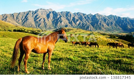 Chestnut horse stands in a sunlit mountain pasture. A distant herd grazes below jagged peaks in warm evening light. 138817345
