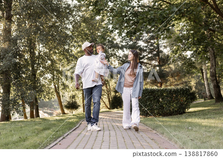 Happy interracial family walking in park on sunny summer day. Father holding little daughter in arms while mother walks. Warm lifestyle, parenting outdoors. People walking Happy interracial family walking in park on sunny summer day. Father holding little daughter in arms while mother walks. Warm lifestyle, parenting outdoors. People walking 138817860
