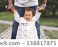 Baby girl taking first steps on park path, African American father's hands holding arms from behind, child milestone candid lifestyle outdoors 138817871