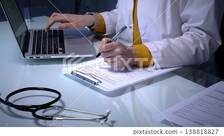 Stethoscope laying on a white reflective desk while medical professional working in clinic office. Typing on laptop and writing notes. Medicine concept 138818827