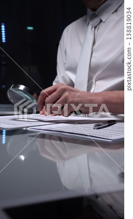 Person examining a document using a magnifying glass, focusing on fine print and terms in a legal agreement, working late. Audit and overworking concepts 138819034