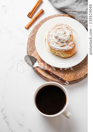 Freshly baked cinnamon roll with white icing and a cup of black coffee on a light marble background. Freshly baked cinnamon roll with white icing and a cup of black coffee on a light marble background. 138819048