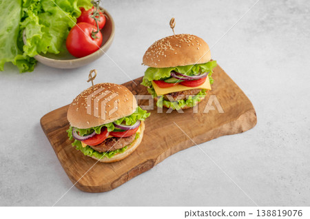 Two beef burgers with tomato, cheese, and lettuce on a cutting board on a light textured background Two beef burgers with tomato, cheese, and lettuce on a cutting board on a light textured background 138819076