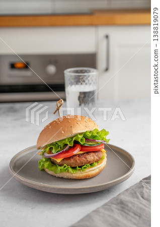 Beef burger with tomato, cheese, and lettuce on a plate on a kitchen table with glass of water Beef burger with tomato, cheese, and lettuce on a plate on a kitchen table with glass of water 138819079