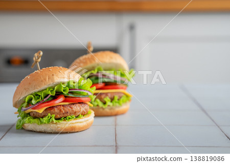 Two meat burgers with tomato, cheese, and lettuce on a table against a white kitchen Two meat burgers with tomato, cheese, and lettuce on a table against a white kitchen 138819086