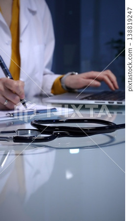 Stethoscope resting on a white reflective desk while medical professional working in clinic office. Wearing lab coat. Typing on laptop and writing notes. 138819247
