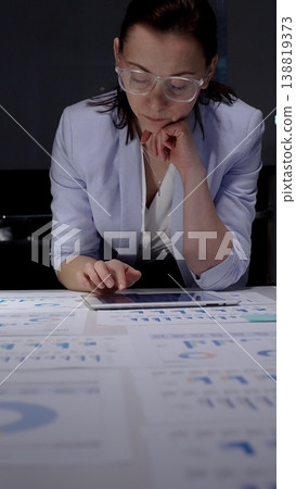 Businesswoman intently reviewing financial charts and reports scattered across her desk while using a digital tablet, working overtime in a corporate environment Businesswoman intently reviewing financial charts and reports scattered across her desk while using a digital tablet, working overtime in a corporate environment 138819373