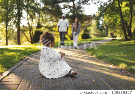 Cute mixed race toddler girl sitting on park path, interracial couple parents walking toward child in sunny green park, candid family lifestyle. Mom, dad and baby Cute mixed race toddler girl sitting on park path, interracial couple parents walking toward child in sunny green park, candid family lifestyle. Mom, dad and baby 138819397