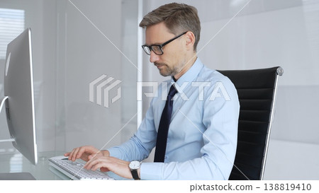 Mature professional businessman in blue shirt and tie focusing on computer screen. Brainstorming ideas and analyzing data. Working on financial strategy in modern corporate office Mature professional businessman in blue shirt and tie focusing on computer screen. Brainstorming ideas and analyzing data. Working on financial strategy in modern corporate office 138819410